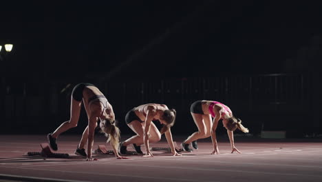 A-row-of-runners-womens-crouch-in-the-starting-position-before-beginning-to-race.-Females-start-with-running-shoes-on-the-stadium-from-the-start-line-in-the-dark-with-spotlights-in-slow-motion.